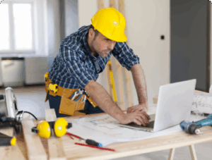 A construction worker wearing a yellow hard hat and tool belt uses a laptop on a workbench with blueprints, tools, and safety gear in a partially finished room.