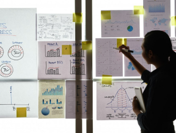 A person stands by a window, analyzing charts and graphs on paper sheets that are attached to the glass with sticky notes.