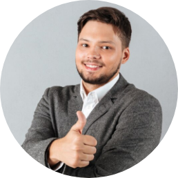 Man wearing a gray suit jacket and white shirt giving a thumbs-up gesture, smiling at the camera against a plain gray background.