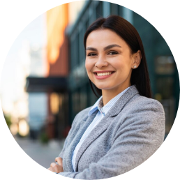 Woman with long dark hair wearing a gray blazer smiles with arms crossed, standing outdoors in an urban setting.