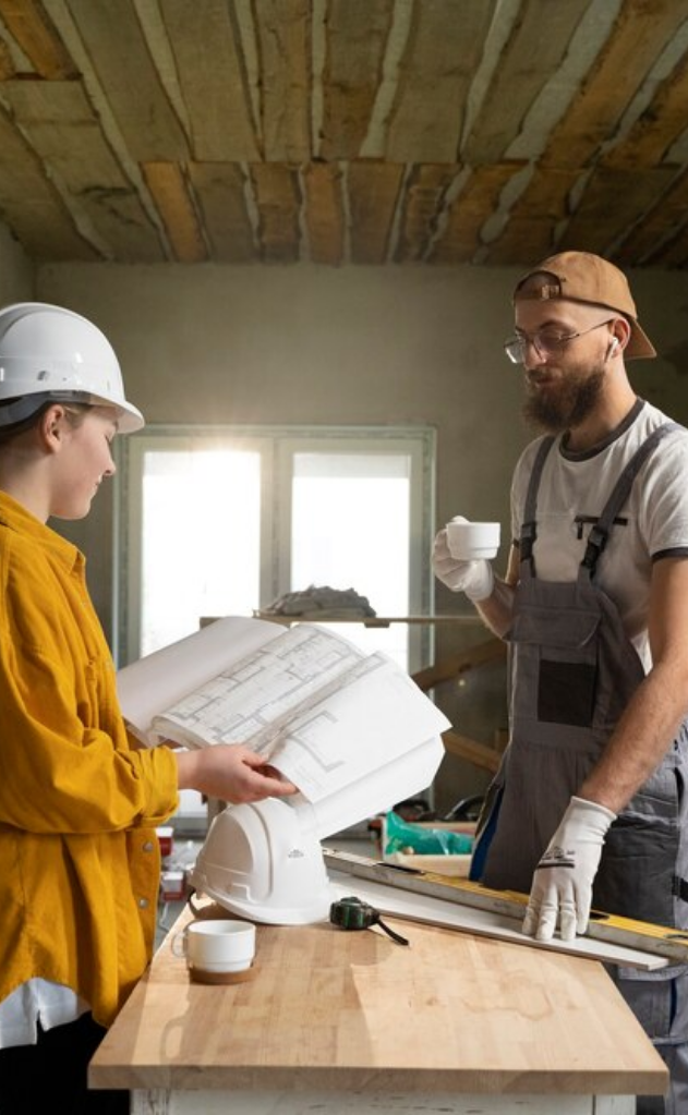 Two construction workers discuss building plans indoors; one holds blueprints while the other drinks coffee. Safety helmets, a tape measure, and tools are on the table.