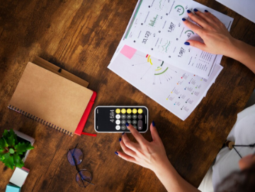 Person reviewing printed charts and graphs while using a calculator on a wooden desk, with a notebook, eyeglasses, and a small potted plant nearby.